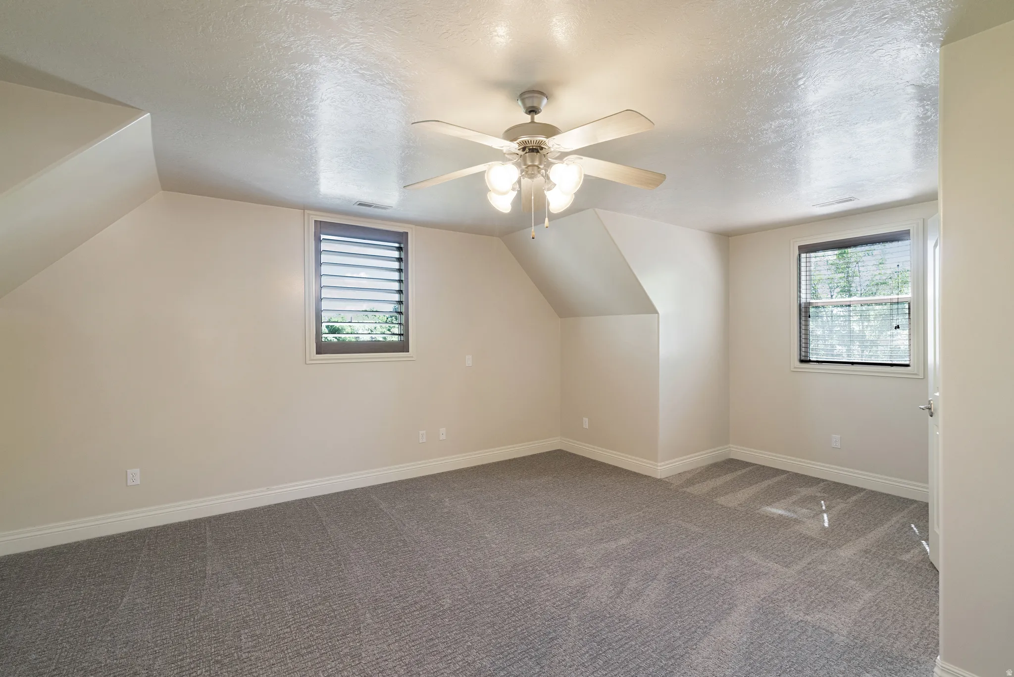 Spacious room featuring vaulted ceilings, a ceiling fan with integrated lighting, two windows for natural light, and plush gray carpeting