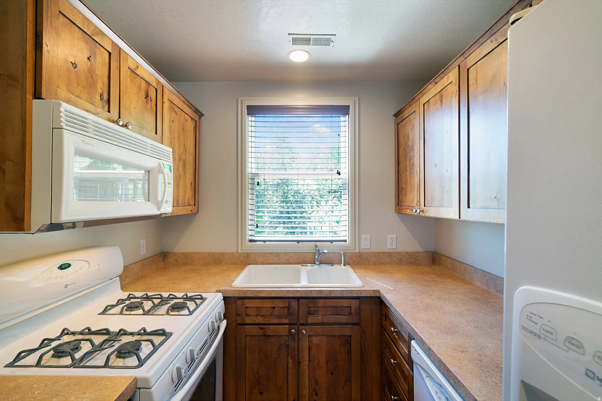 U-shaped kitchen featuring wood cabinetry, laminate countertops, a double basin sink with window, a gas range with an over-the-range microwave, and a built-in dishwasher