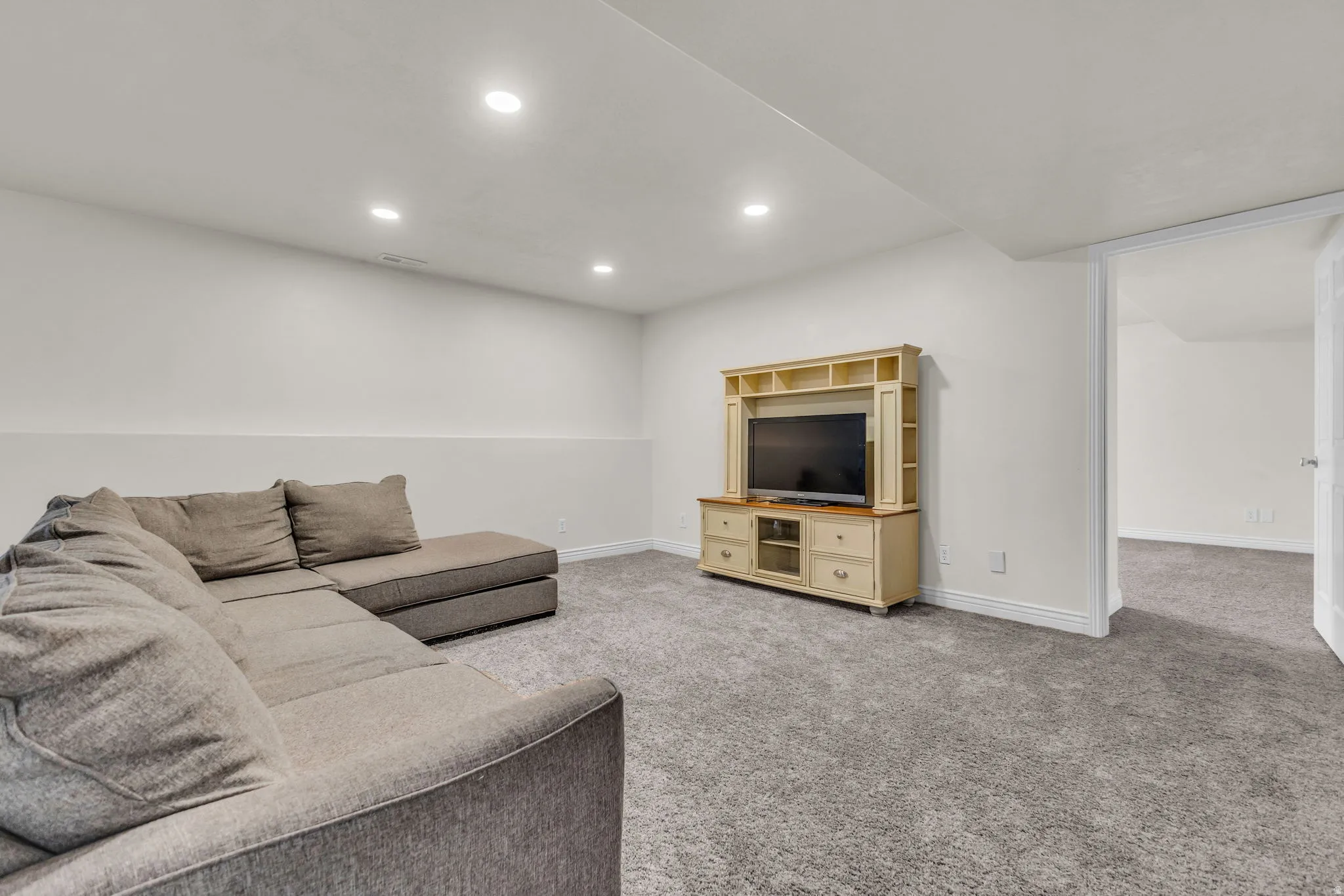 Recessed ceiling lighting illuminates a spacious room featuring light gray carpet flooring and white painted walls