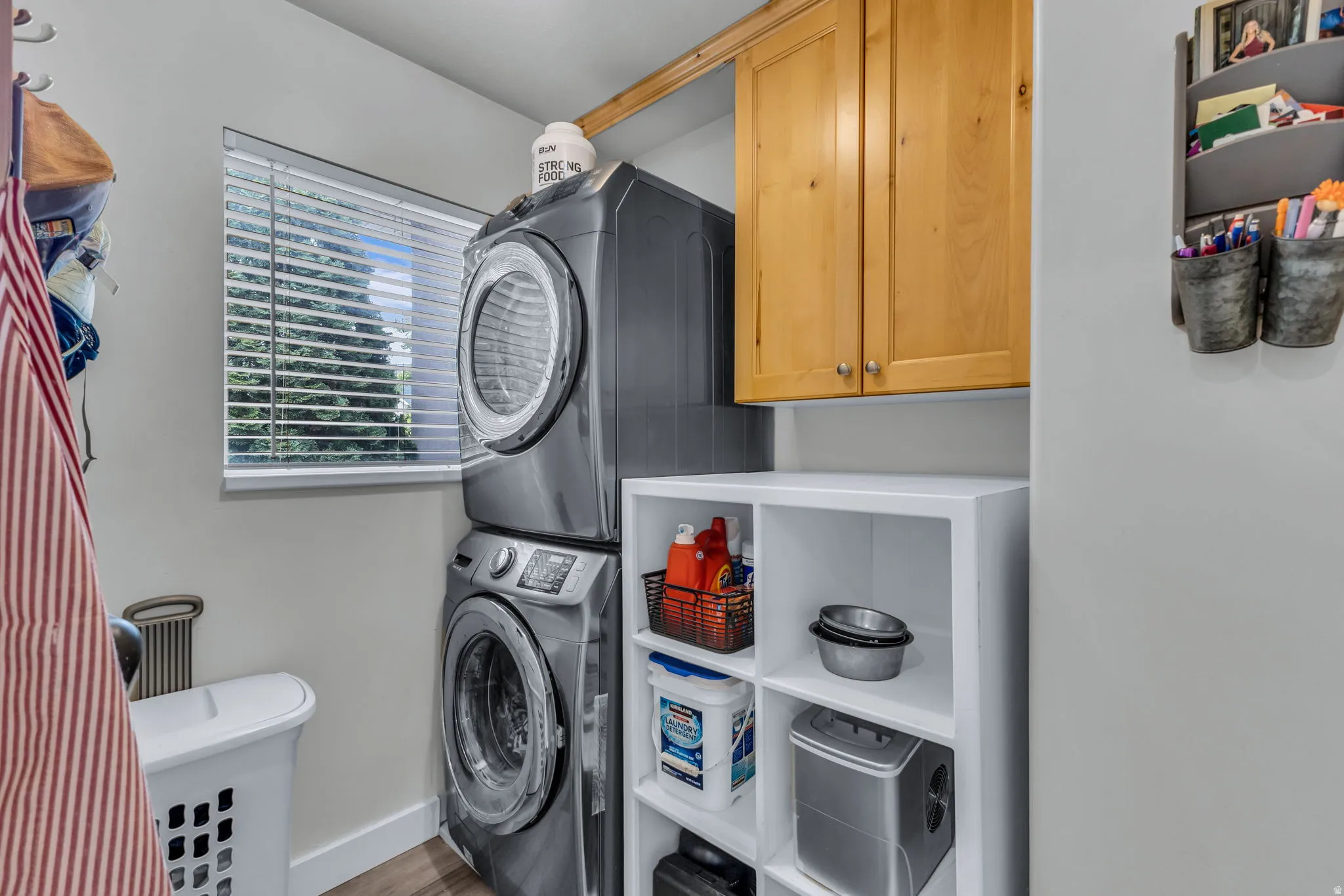 Dedicated laundry area featuring modern stacked appliances, a window with horizontal blinds, built-in natural wood cabinetry, and open shelving storage