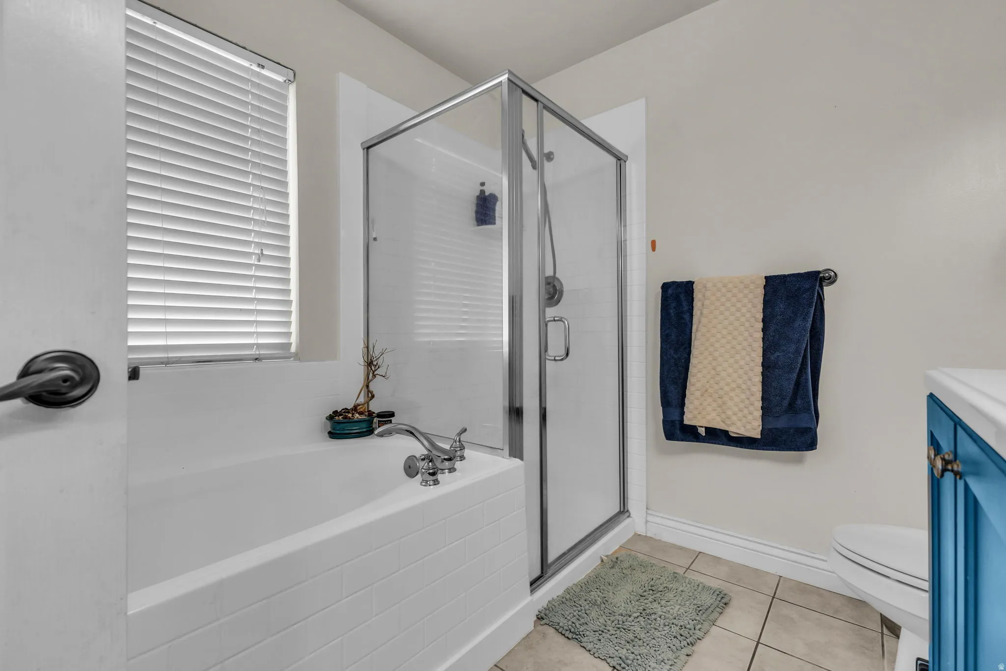 Bathroom featuring a white bathtub, a clear glass shower enclosure, beige floor tiles, and a window with white blinds