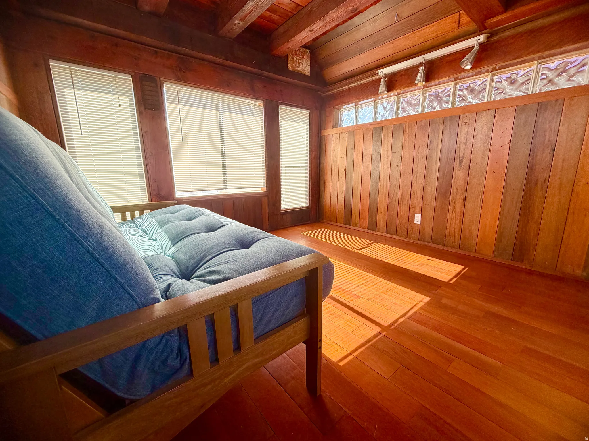 Wood-paneled room featuring wood-finish flooring, exposed beam ceiling, horizontal window blinds, upper glass block accents, and track lighting