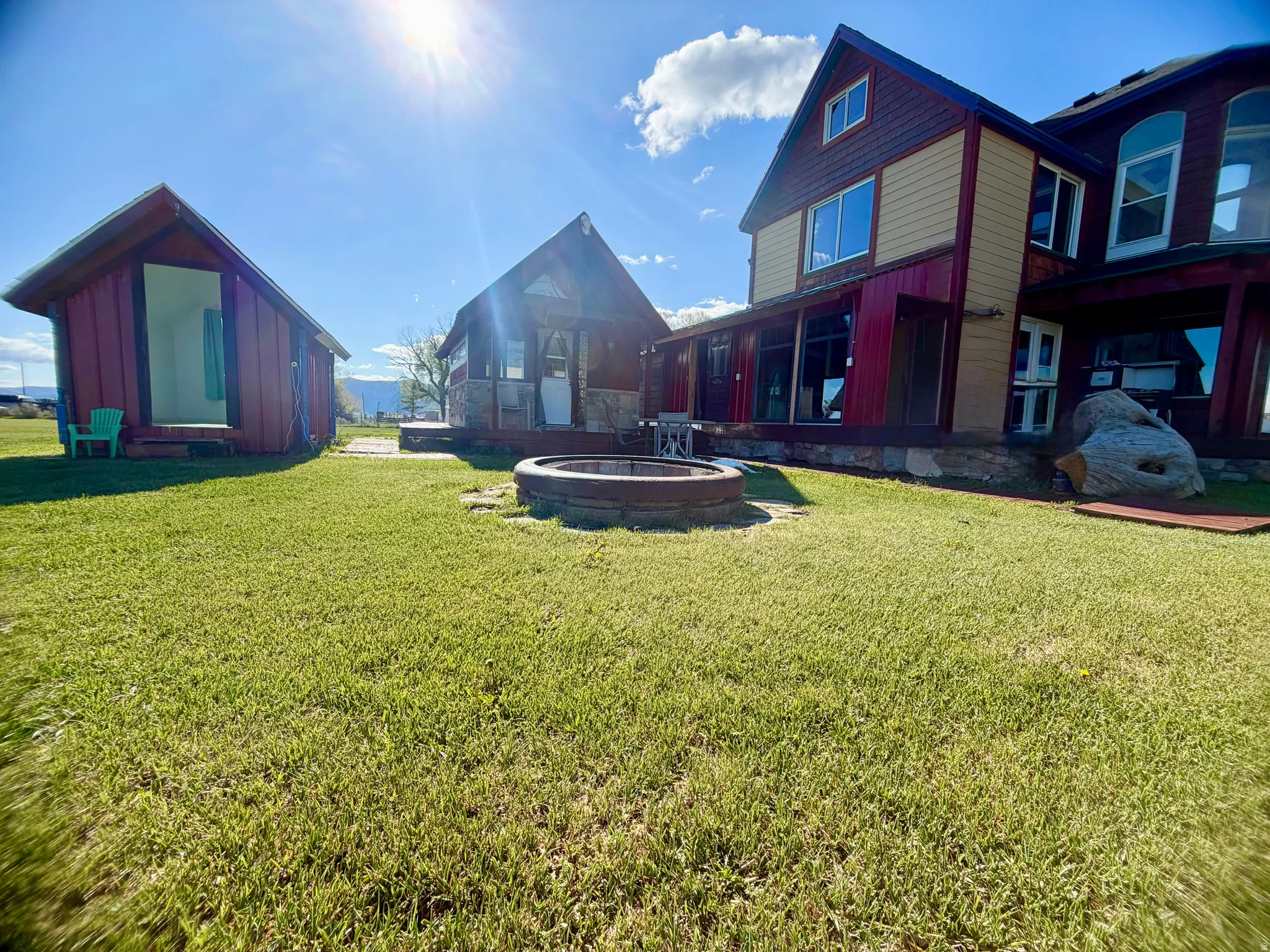Expansive lawn area featuring a central fire pit, two separate outbuildings with red siding, and a main residence with a covered porch