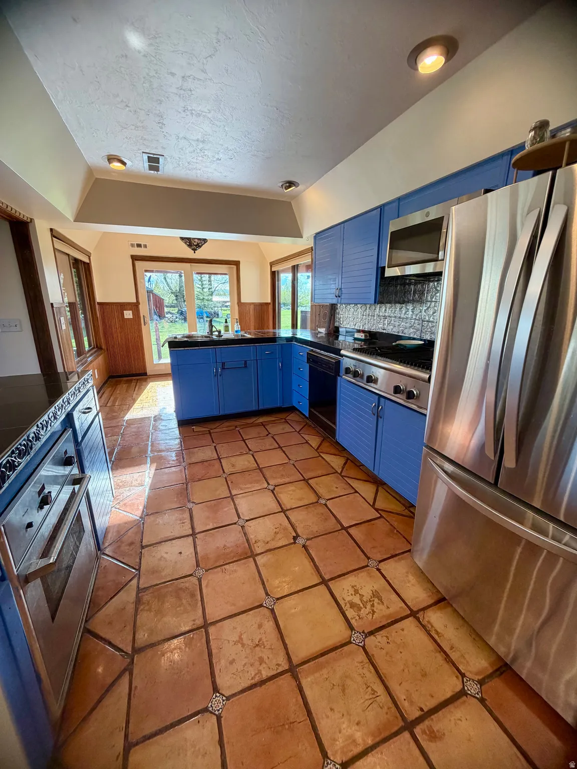 Kitchen featuring terracotta tile flooring, blue cabinetry, stainless steel appliances, dark countertops, and a tiled backsplash