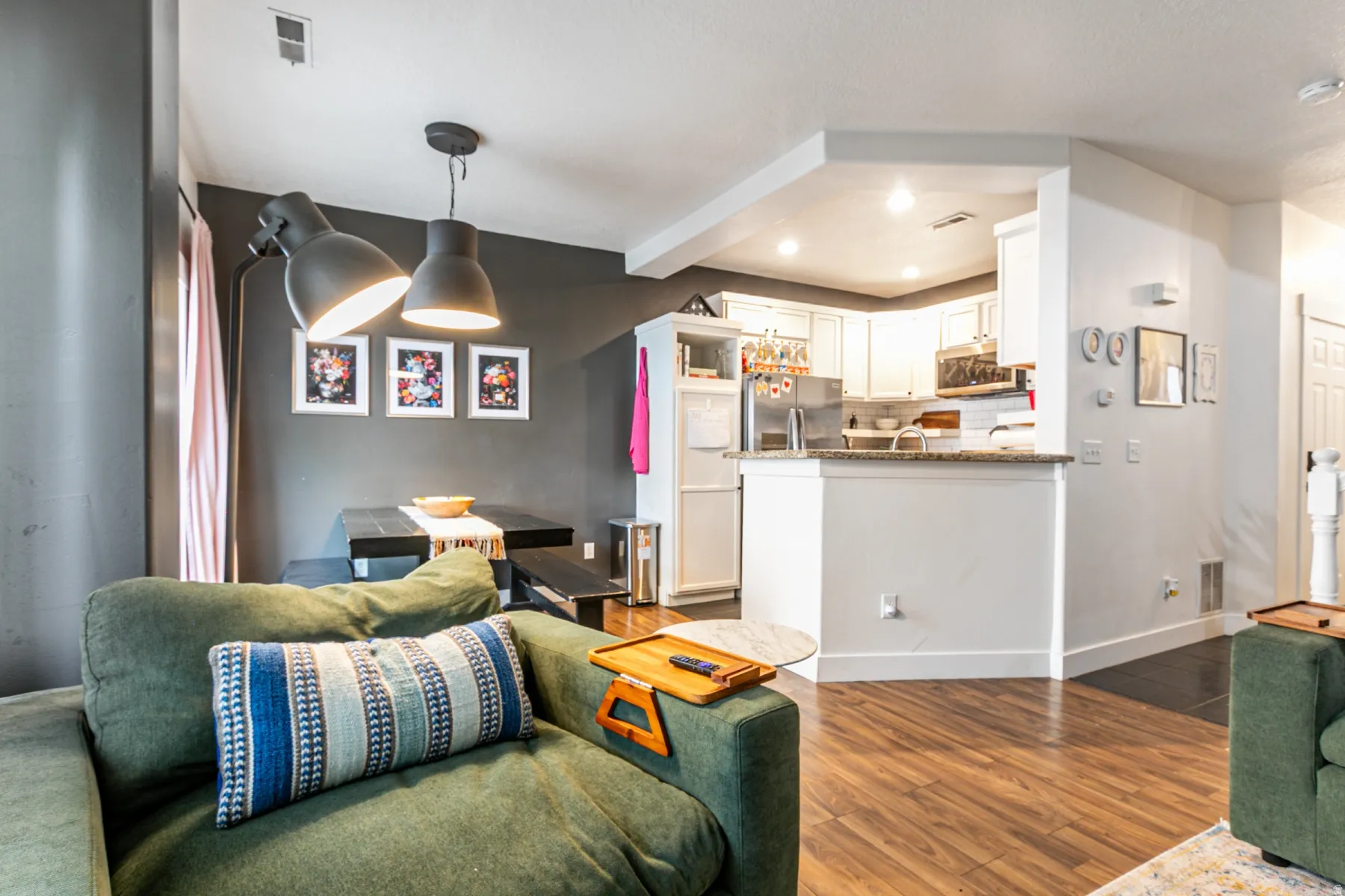 Open-concept living area featuring wood-finish flooring, a contemporary pendant light fixture, and a kitchen with white cabinetry, stainless steel appliances, and a granite-style countertop