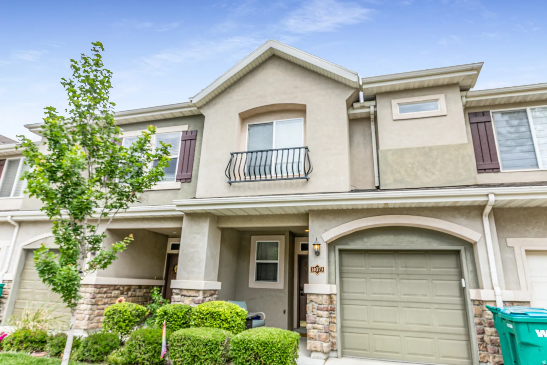 Stucco exterior featuring stone veneer accents, an arched garage entrance, a Juliet balcony with an iron railing, decorative window shutters, and manicured shrubbery