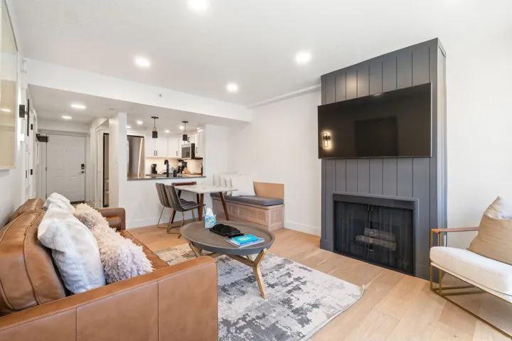 Living area featuring a modern fireplace with a dark paneled surround, light wood-finish flooring, recessed lighting, and a partial view of the kitchen