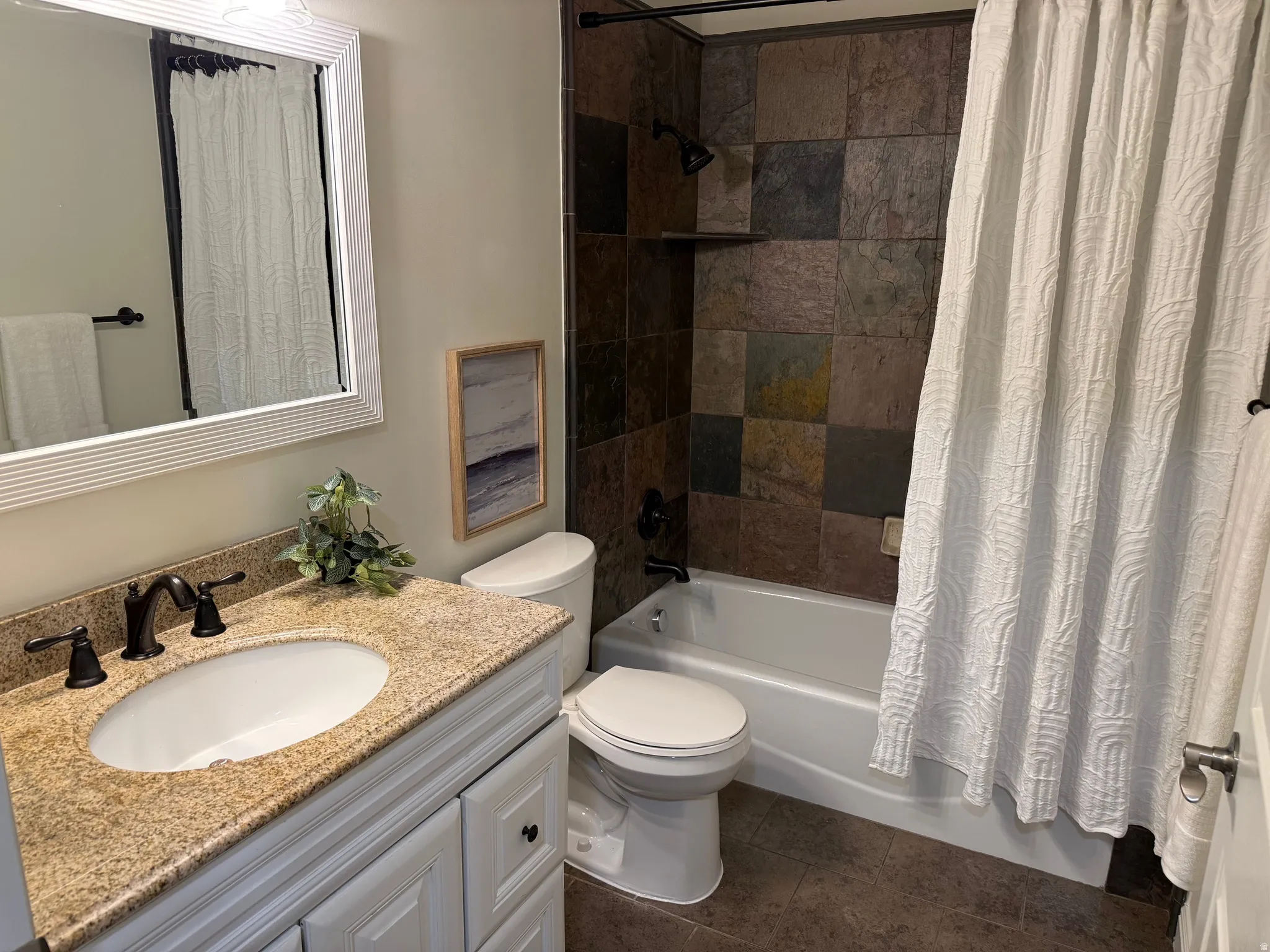 Bathroom featuring a vanity with a light speckled countertop and undermount sink, dark bronze-finish fixtures, a white framed mirror, and a tiled shower surround