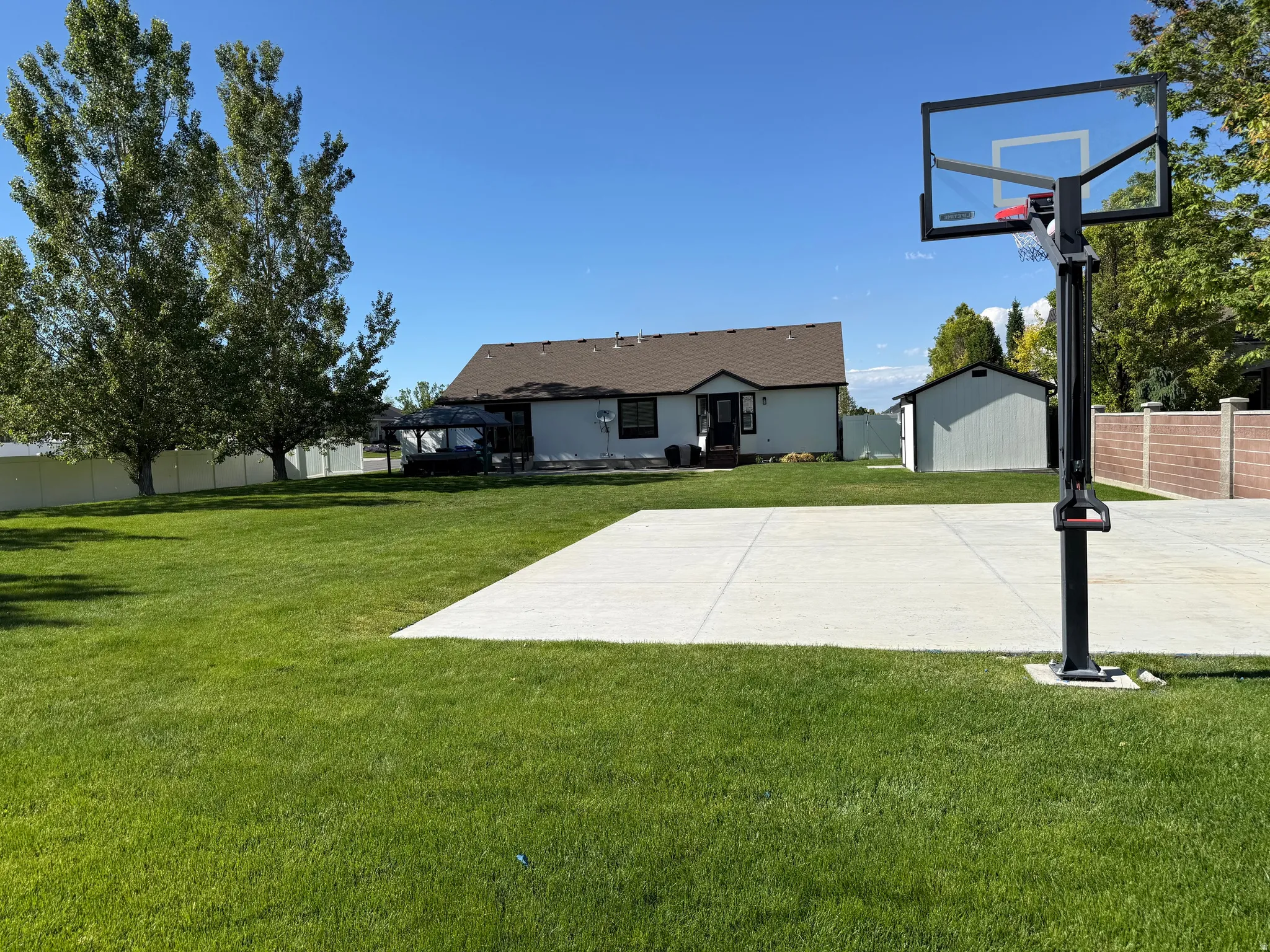 Expansive backyard featuring a large concrete pad, mature trees, a white vinyl privacy fence, and a storage shed