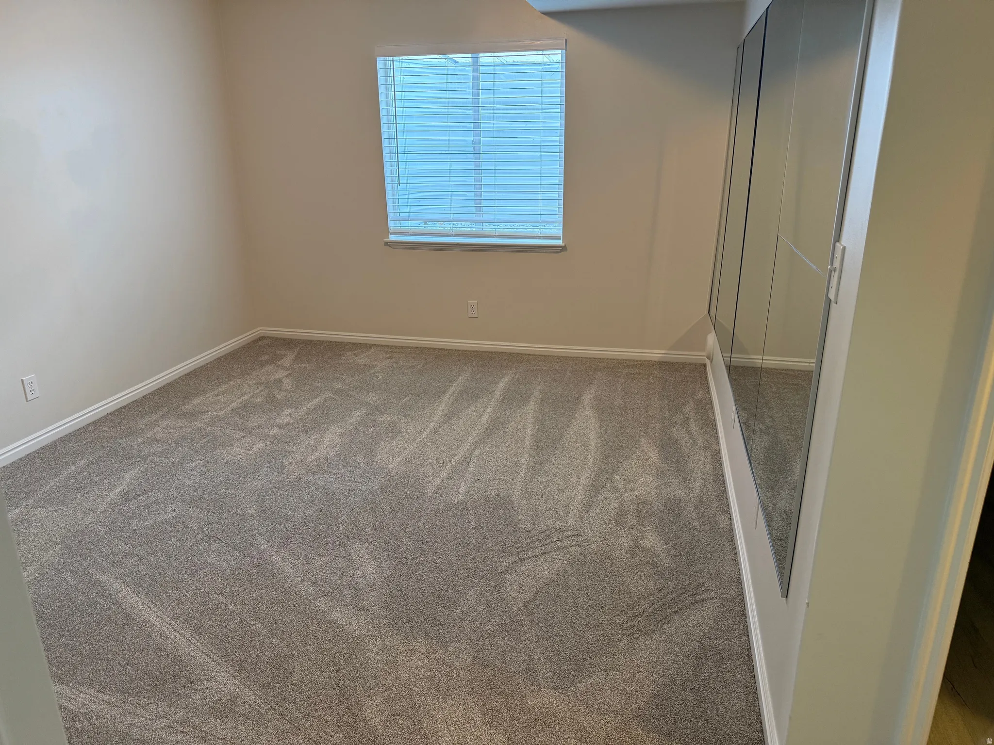 Room featuring soft gray carpeting, a window with blinds, and mirrored sliding closet doors