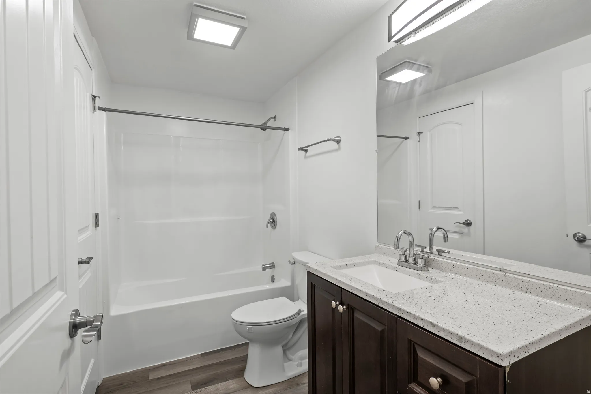 Bathroom featuring a white tub/shower combination, dark wood-finish vanity with a light speckled countertop, and wood-finish flooring