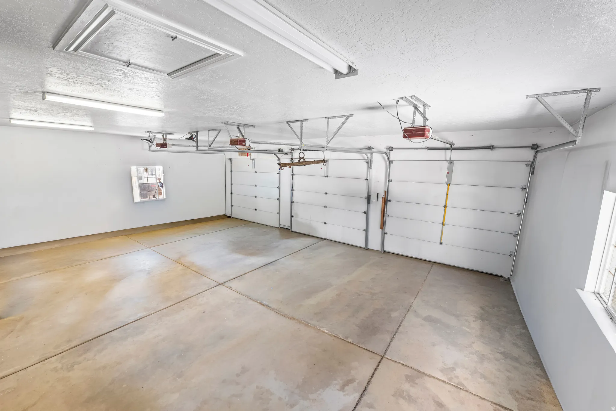 Spacious garage featuring three individual garage doors with automatic openers, a durable concrete floor, bright overhead fluorescent lighting, and an attic access panel