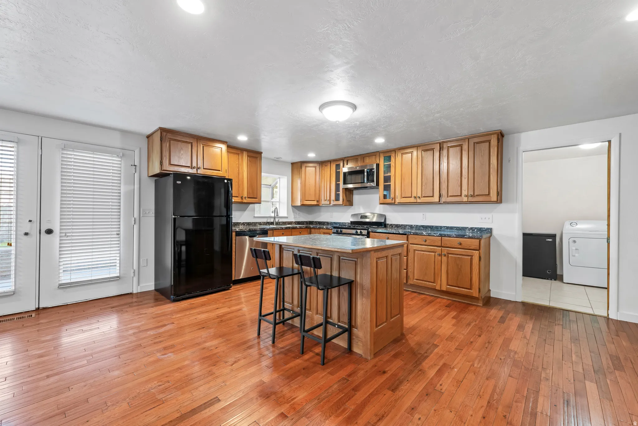 Kitchen featuring wood-finish flooring, a central island with seating, wood cabinetry, stainless steel appliances including a microwave, and a black refrigerator
