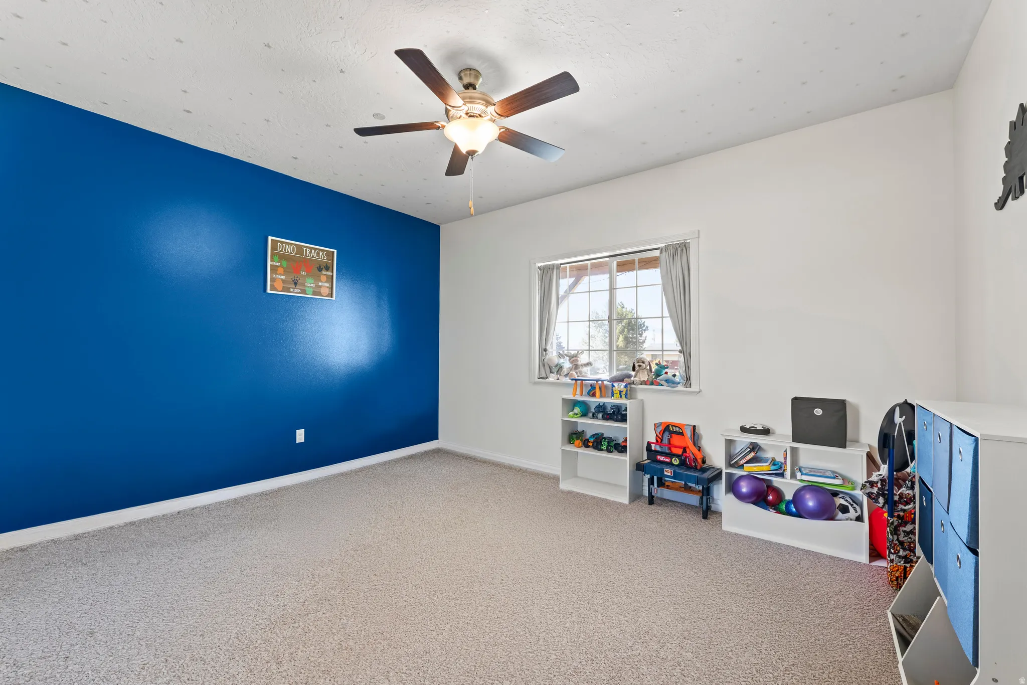 Carpeted room featuring a ceiling fan with light fixture, a window with a wood frame, and a contrasting accent wall