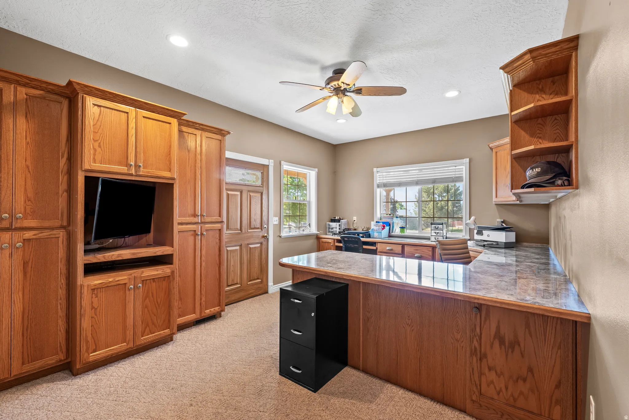 Dedicated office space featuring extensive built-in wood cabinetry, a substantial L-shaped desk with a stone-finish countertop, recessed lighting, and a ceiling fan