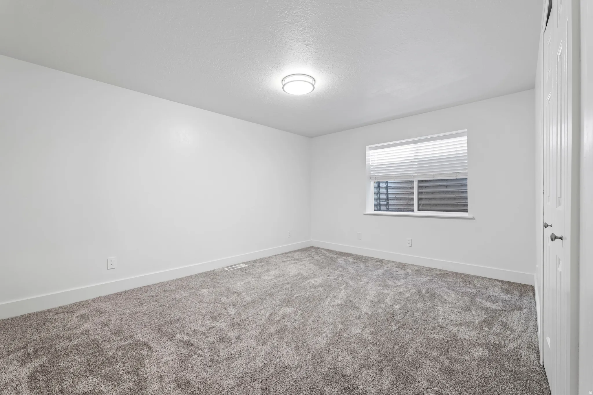 Spacious room featuring neutral carpeting, white walls, a single window with blinds, and a flush-mount ceiling light fixture