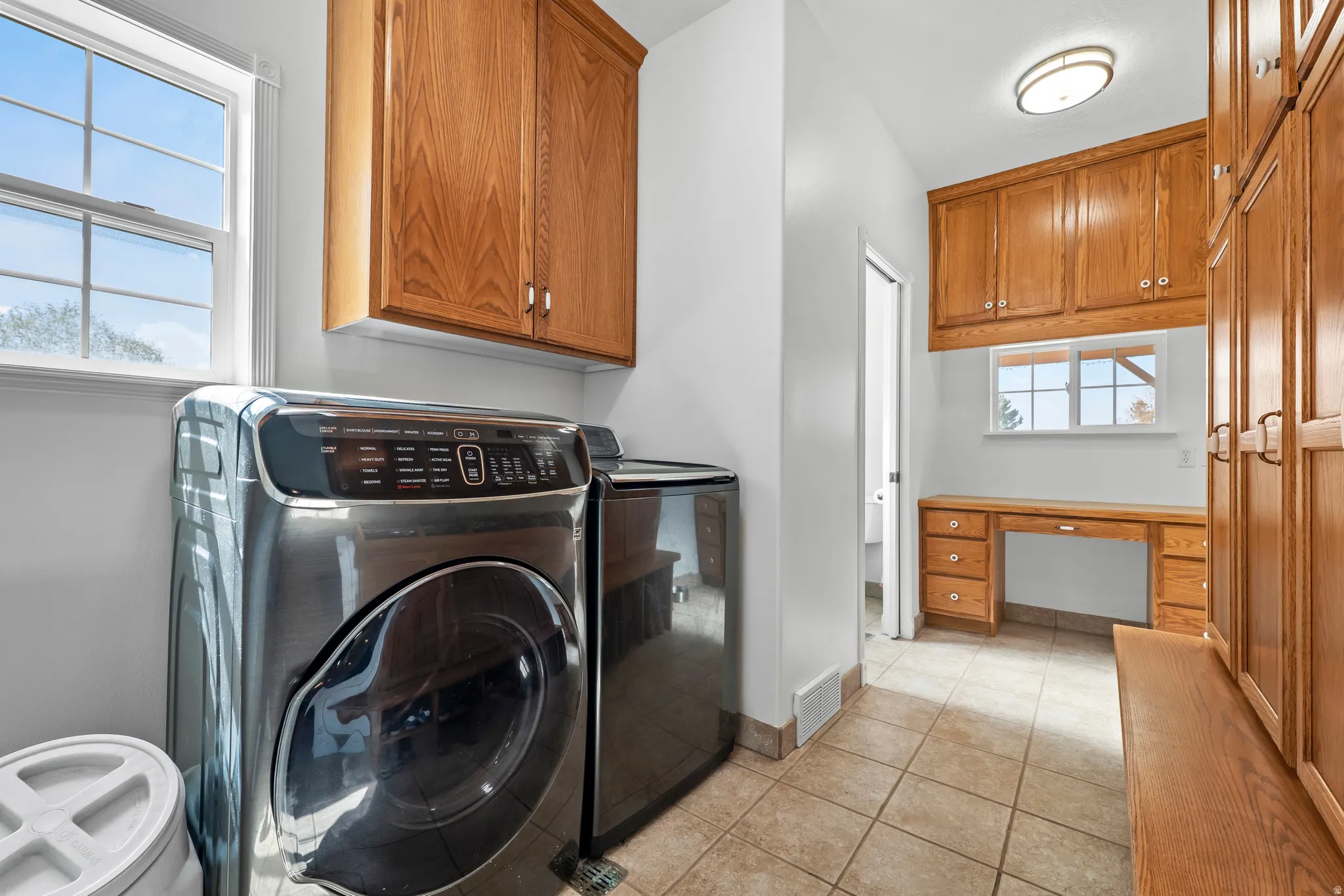 Functional laundry area featuring a front-load washer, natural light from a double-pane window, extensive built-in cabinetry, an integrated desk space, and neutral tile flooring