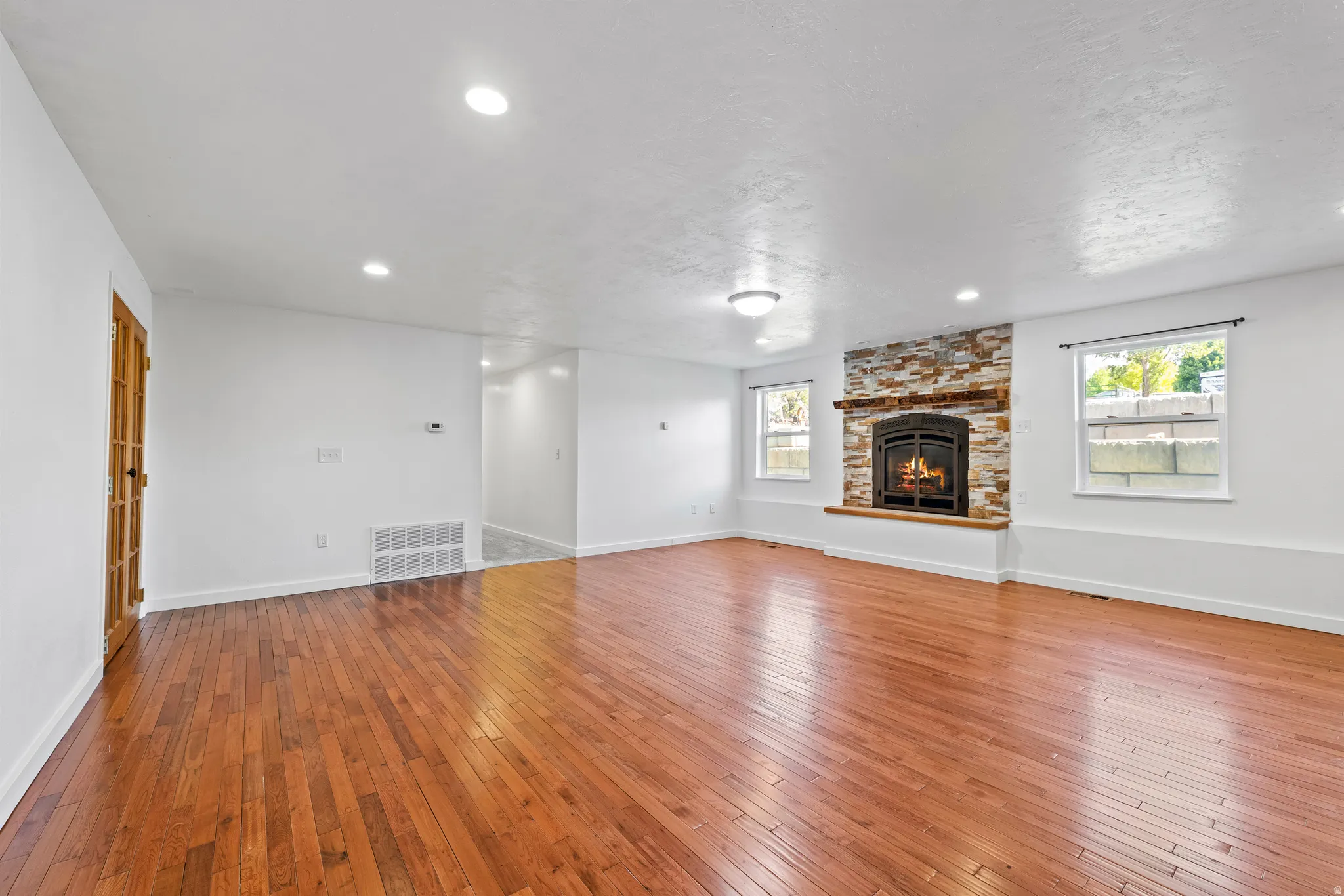 Spacious living area featuring warm wood-finish flooring, a stone-clad fireplace with a decorative mantel, multiple windows, and recessed ceiling lighting