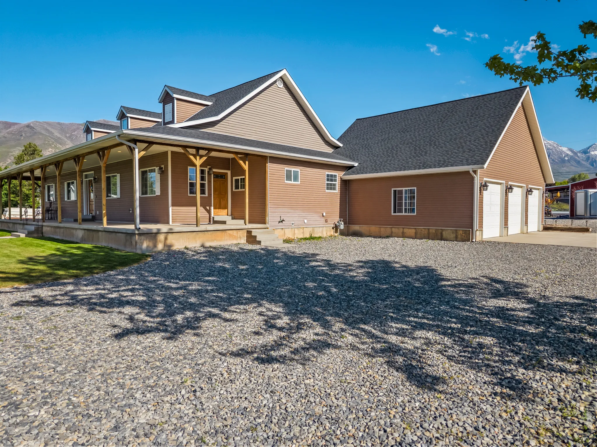 Expansive gravel driveway leading to a residence featuring a wrap-around porch with natural wood pillars, dormer windows, and a three-car garage