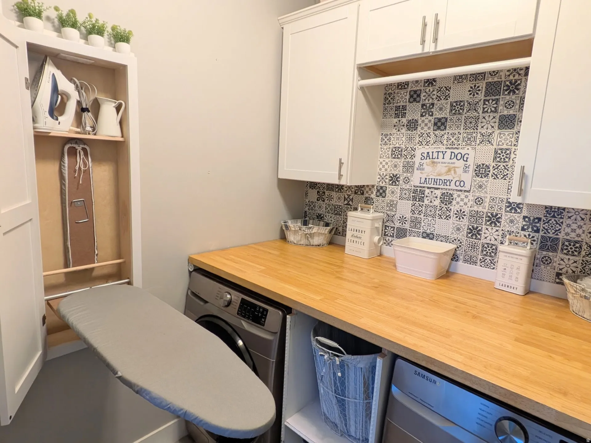 Dedicated laundry space featuring a built-in ironing board cabinet, white upper cabinetry, and a wood-finish countertop