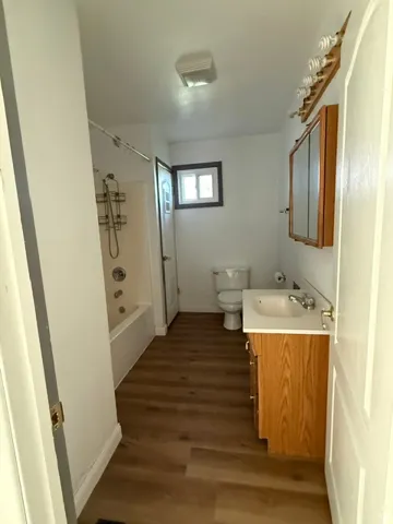 UPPER LEVEL BATHROOM- Wood-finish flooring, white bathtub and shower surround, light wood-tone vanity with white countertop, white toilet, and recessed window with dark trim