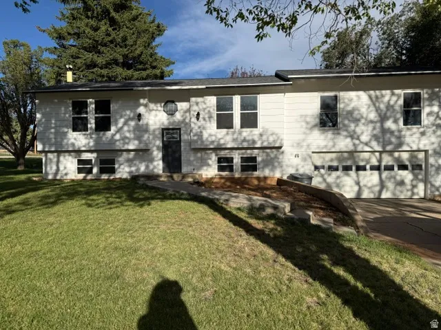 White exterior with a two-car garage, mature trees, a sloped front lawn, and a paved driveway
