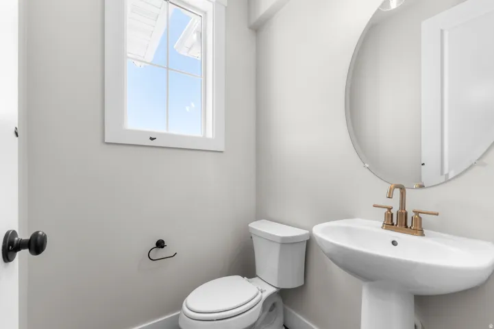 Pedestal sink with brass-finish faucet, circular frameless mirror, and a window with white trim