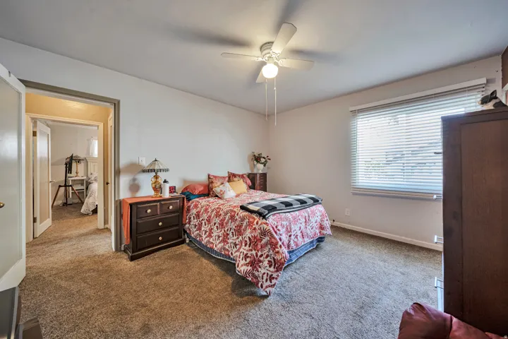 Carpeted bedroom featuring a ceiling fan with integrated lighting, a window with horizontal blinds, and a doorway leading to an additional space