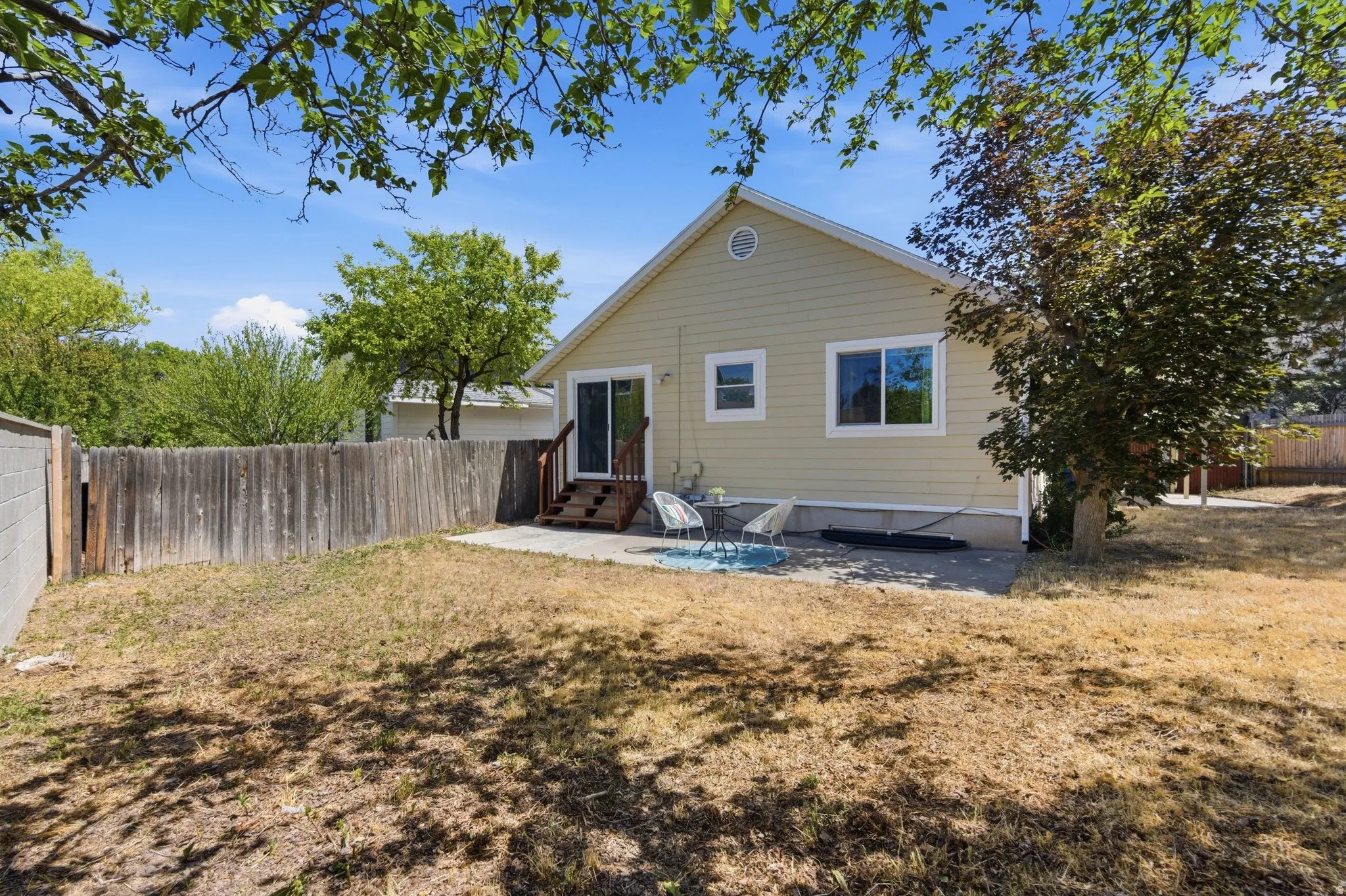 Rear exterior featuring a sliding glass door, patio area, wood fencing, mature trees, and a pitched roofline