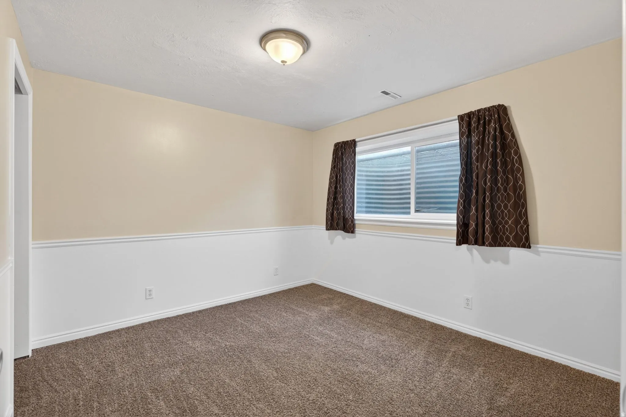 Carpeted room featuring a neutral two-tone wall finish with wainscoting, an egress window, and a flush-mount ceiling light fixture