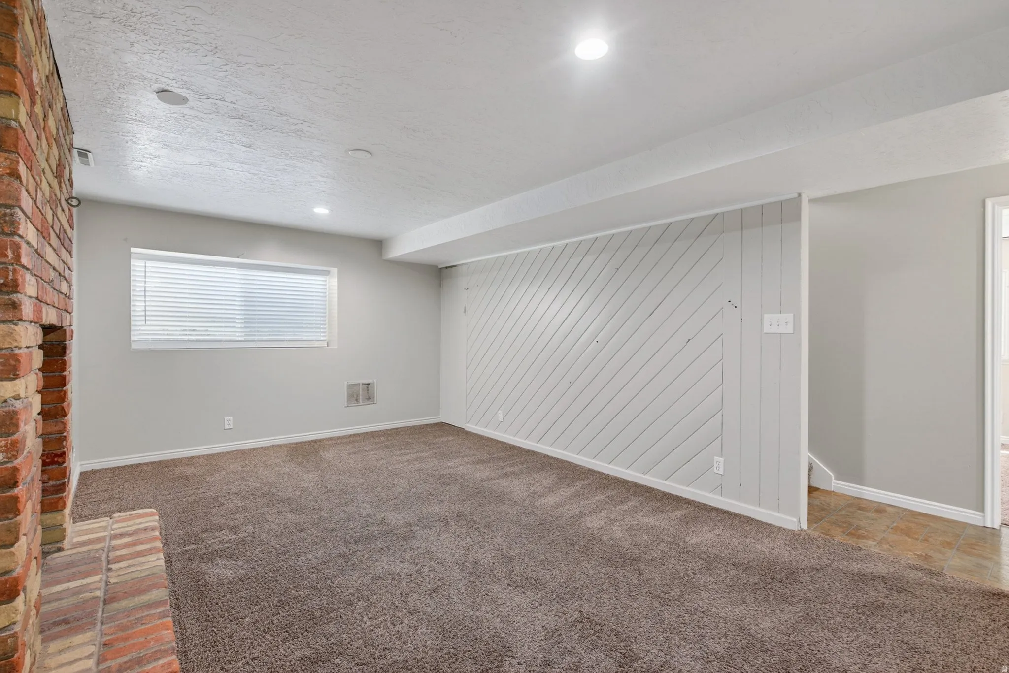 Spacious room featuring a brick fireplace, neutral carpeting, light gray walls, a window with blinds, and white diagonal shiplap detailing