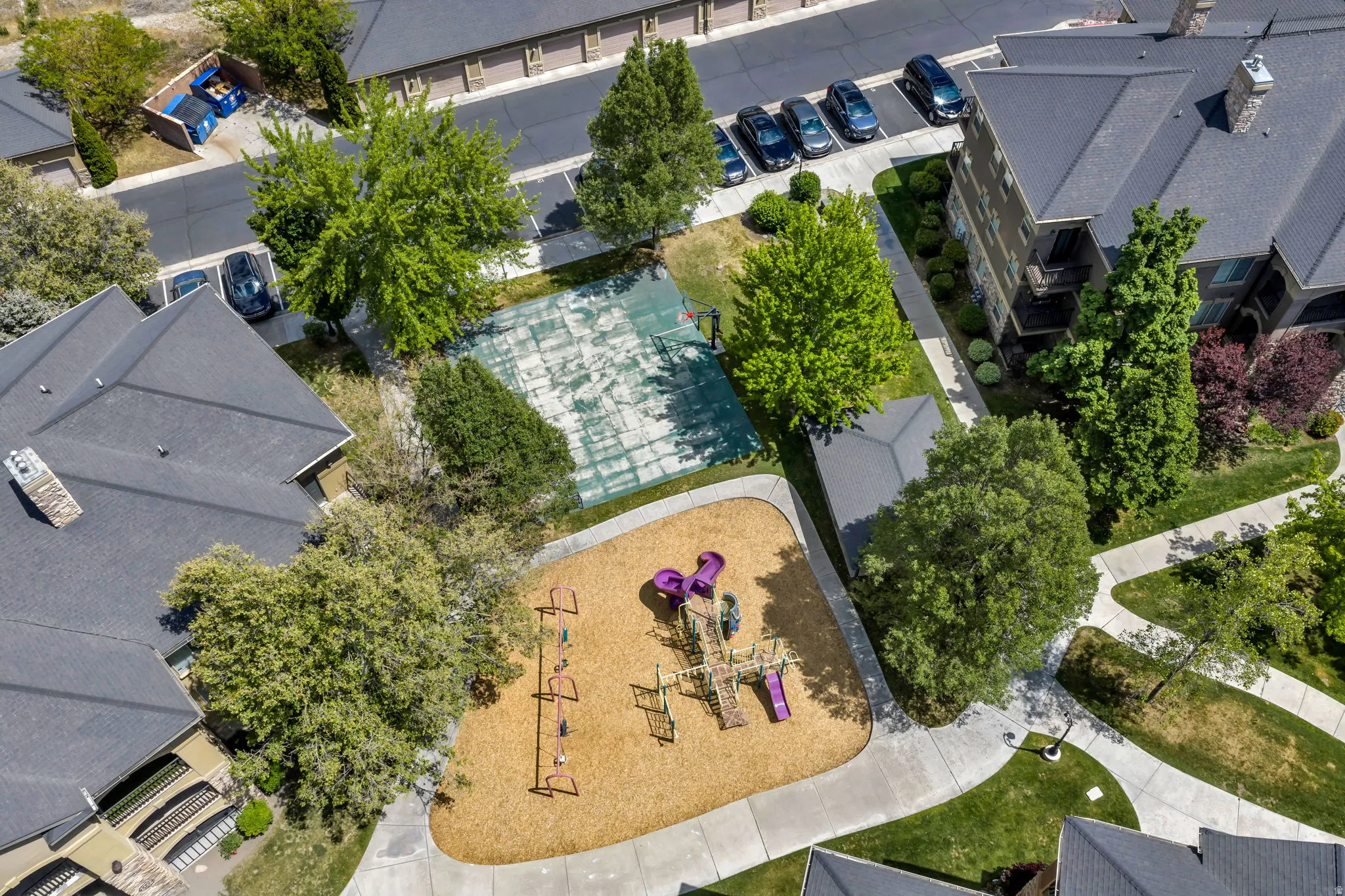 Outdoor recreation area featuring a playground with wood chip surfacing, a multi-sport court, landscaped green spaces, mature trees, and paved pedestrian pathways