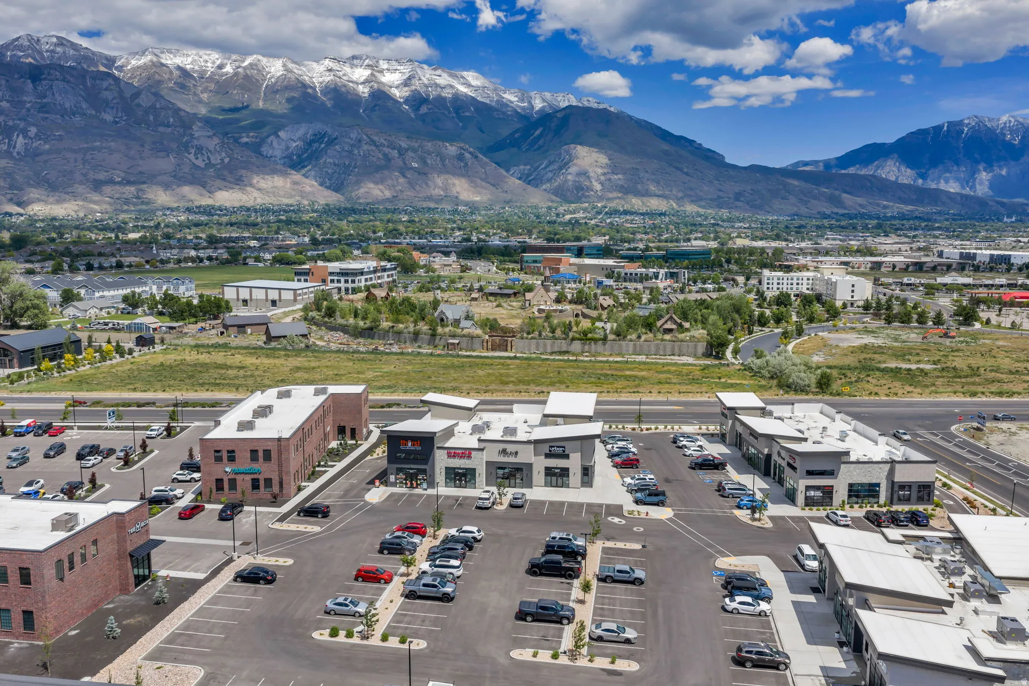 Commercial development featuring multiple buildings, expansive paved parking, and a prominent mountainous backdrop