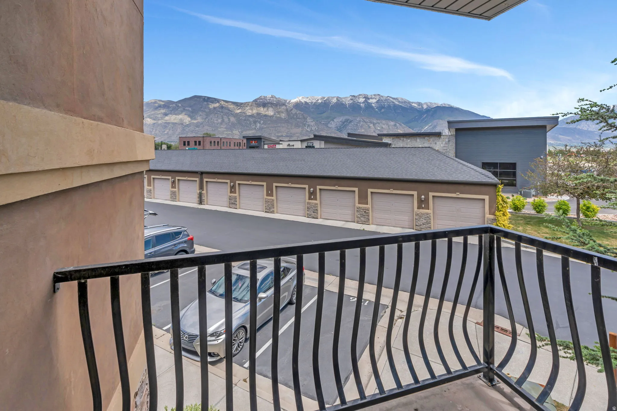 Balcony with decorative iron railing overlooking an asphalt parking area and a row of garages with stone accents