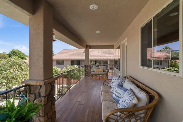 Covered outdoor balcony featuring wood-finish flooring, stucco columns with stone bases, black wrought iron railing, and multiple windows