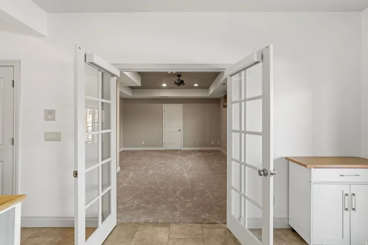 Double French doors open to a spacious room featuring a tray ceiling with recessed lighting and a ceiling fan, plush carpeting, and light gray wall paint