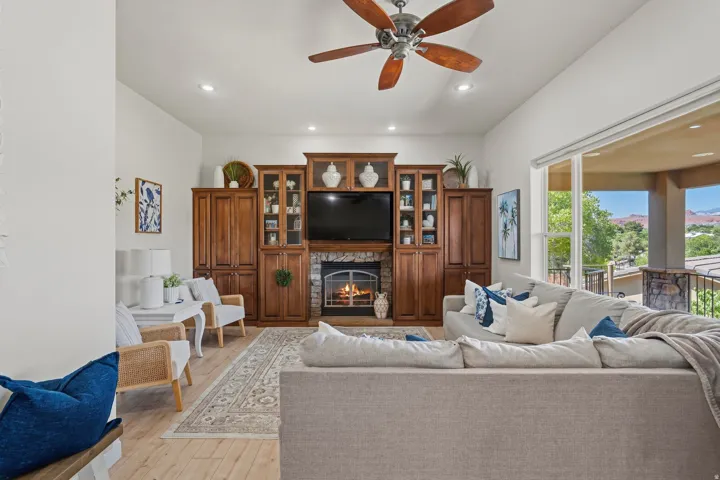 Living area featuring a stone-surround fireplace with a hearth, custom built-in cabinetry, wood-finish flooring, and a ceiling fan