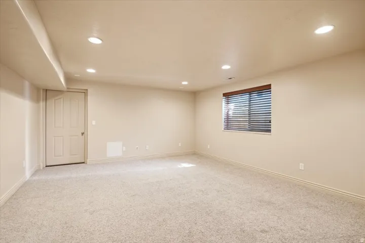 Spacious room featuring neutral carpeting, a single window with horizontal blinds, recessed ceiling lighting, and a white paneled door