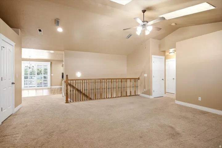Spacious carpeted area featuring a wood staircase with iron balusters, vaulted ceilings with skylights, and a ceiling fan with integrated lighting