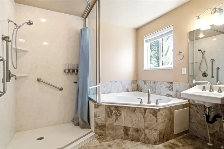 Bathroom featuring a tiled corner soaking tub, walk-in shower with grab bars and built-in shelving, pedestal sink with wall-mounted mirror, and overhead vanity lighting