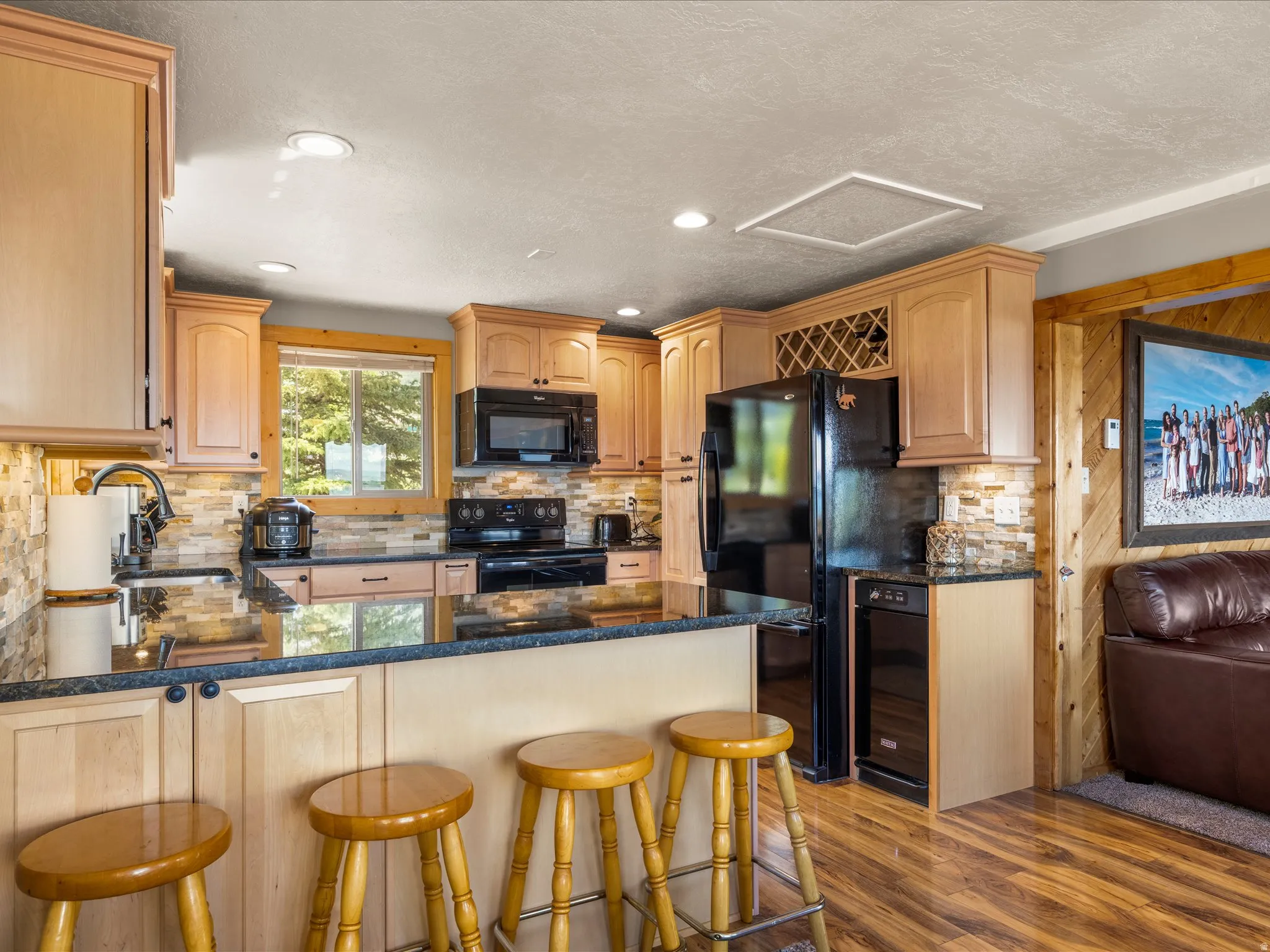 Kitchen featuring light wood cabinetry, black appliances, a stone tile backsplash, dark countertops, and wood-finish flooring