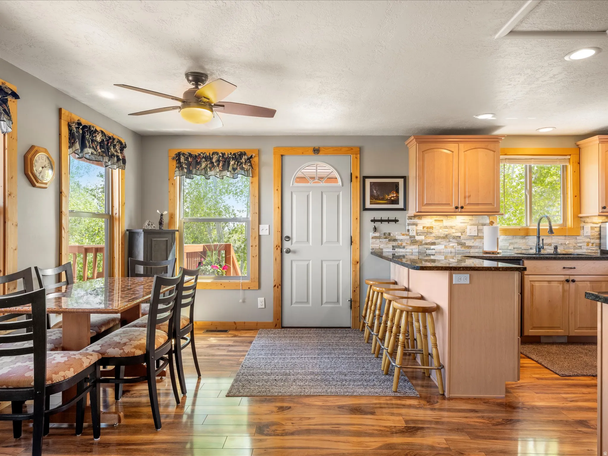 Open-concept kitchen and dining area featuring wood-finish flooring, light wood cabinetry, a granite countertop island with seating, a stone tile backsplash, and multiple windows with wood trim