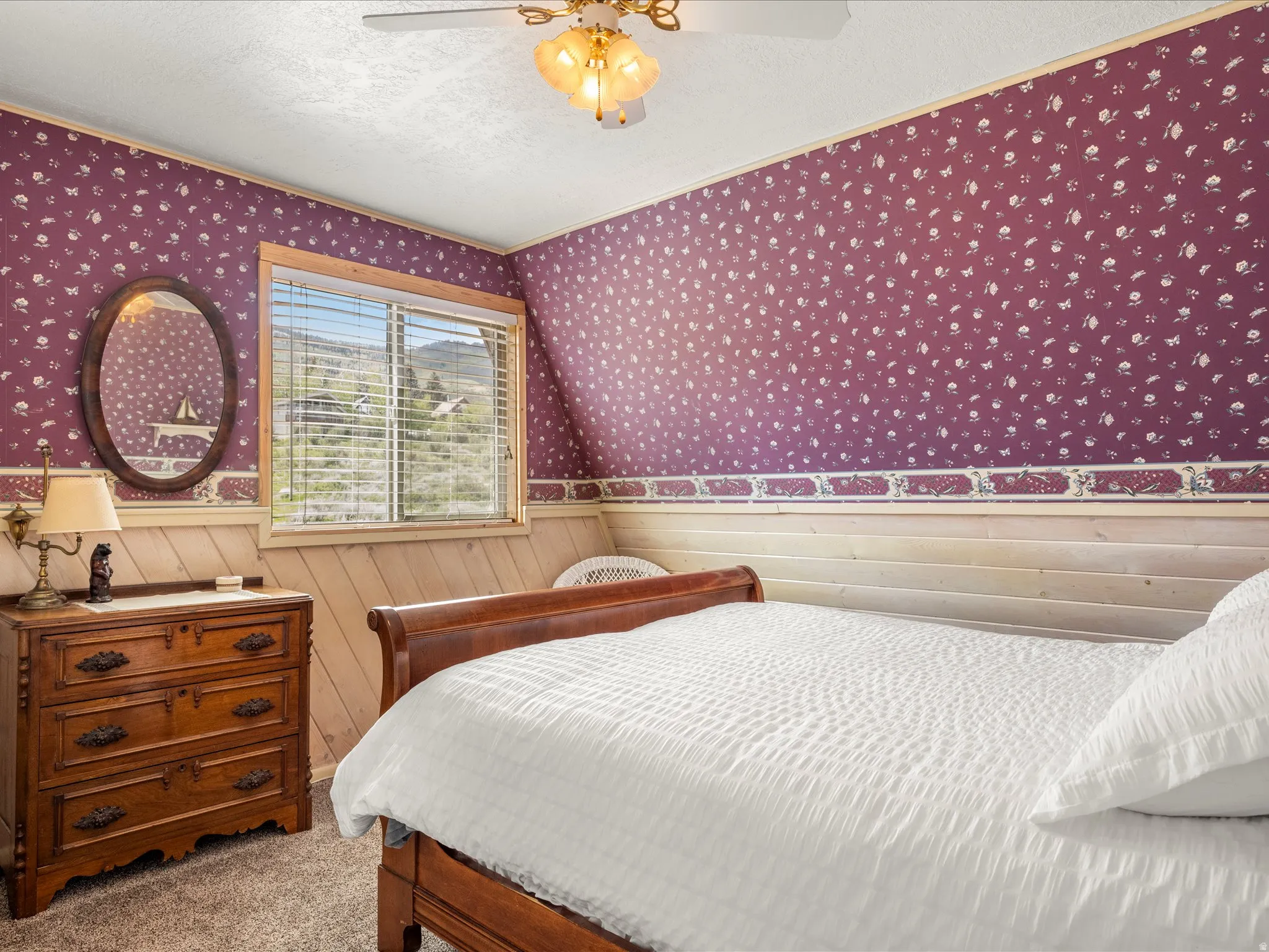 Bedroom featuring a window with integrated blinds, patterned wallpaper, natural wood paneling, and a ceiling fan with integrated lighting