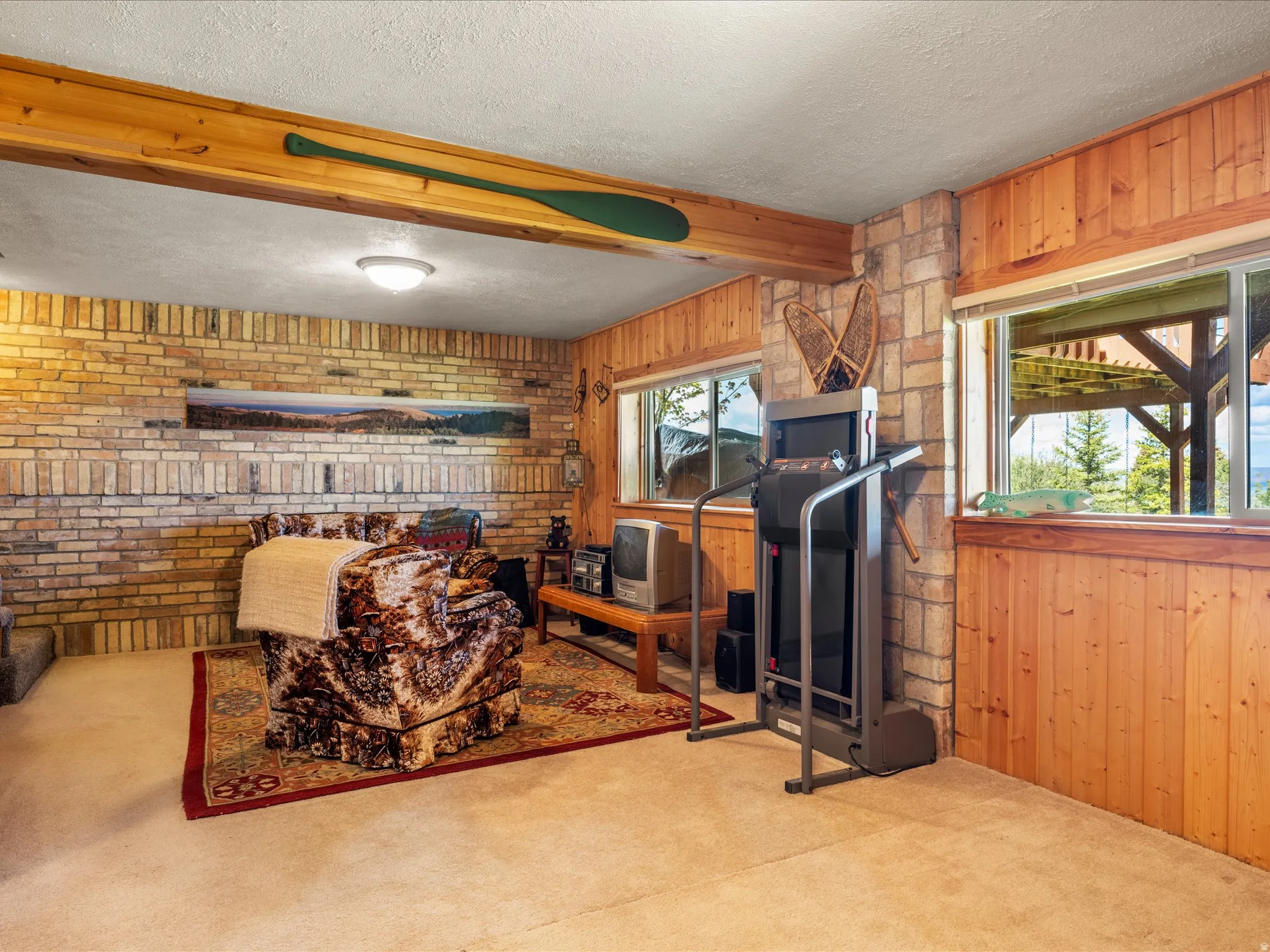 Carpeted room featuring brick accent walls, wood paneling, wood-finish ceiling beam, and windows overlooking exterior scenery