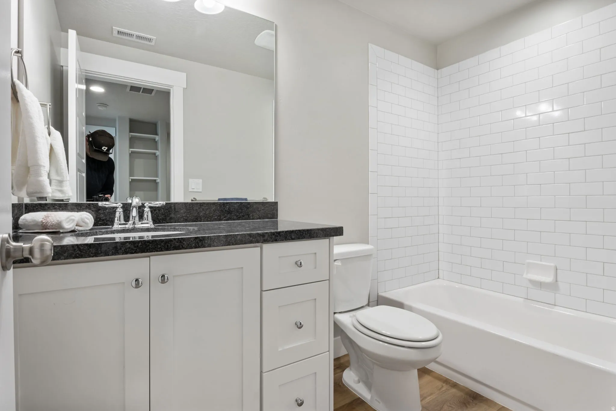 Bathroom featuring a white subway tile surround bathtub, a single vanity with a dark countertop, an undermount sink, and wood-finish flooring