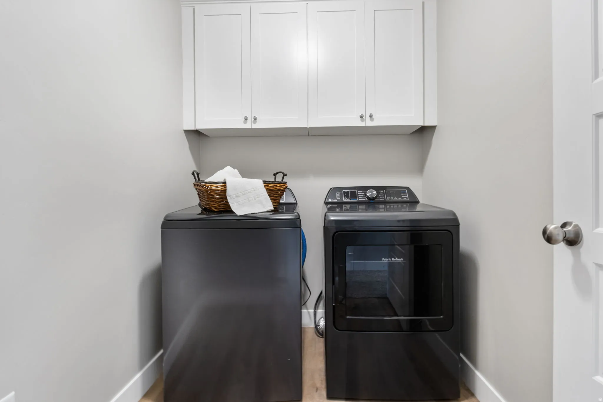 Dedicated laundry area featuring overhead white cabinetry with brushed nickel hardware, light gray walls, and wood-finish flooring