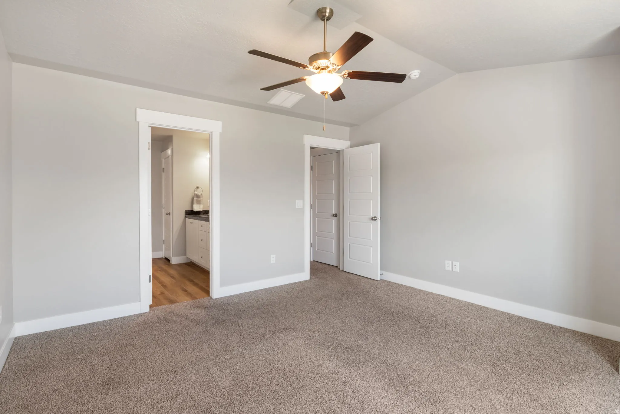 Carpeted interior space featuring a ceiling fan with integrated lighting, a vaulted ceiling, and an entryway to a bathroom with wood-finish flooring, a vanity, and a wall-mounted light fixture