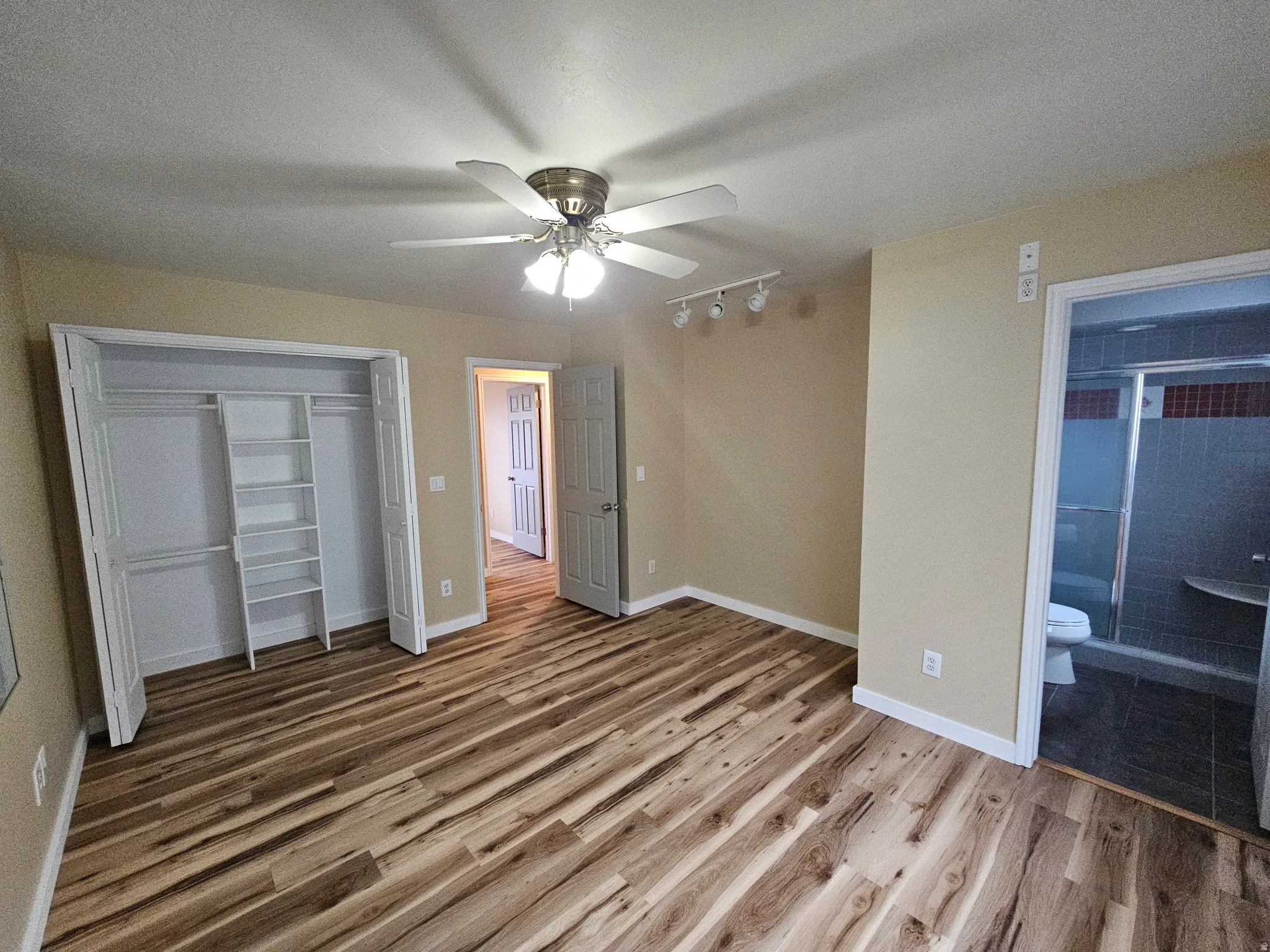 Spacious room featuring wood-finish flooring, a ceiling fan with integrated lighting, track lighting fixtures, and a built-in closet system with shelving and hanging rods