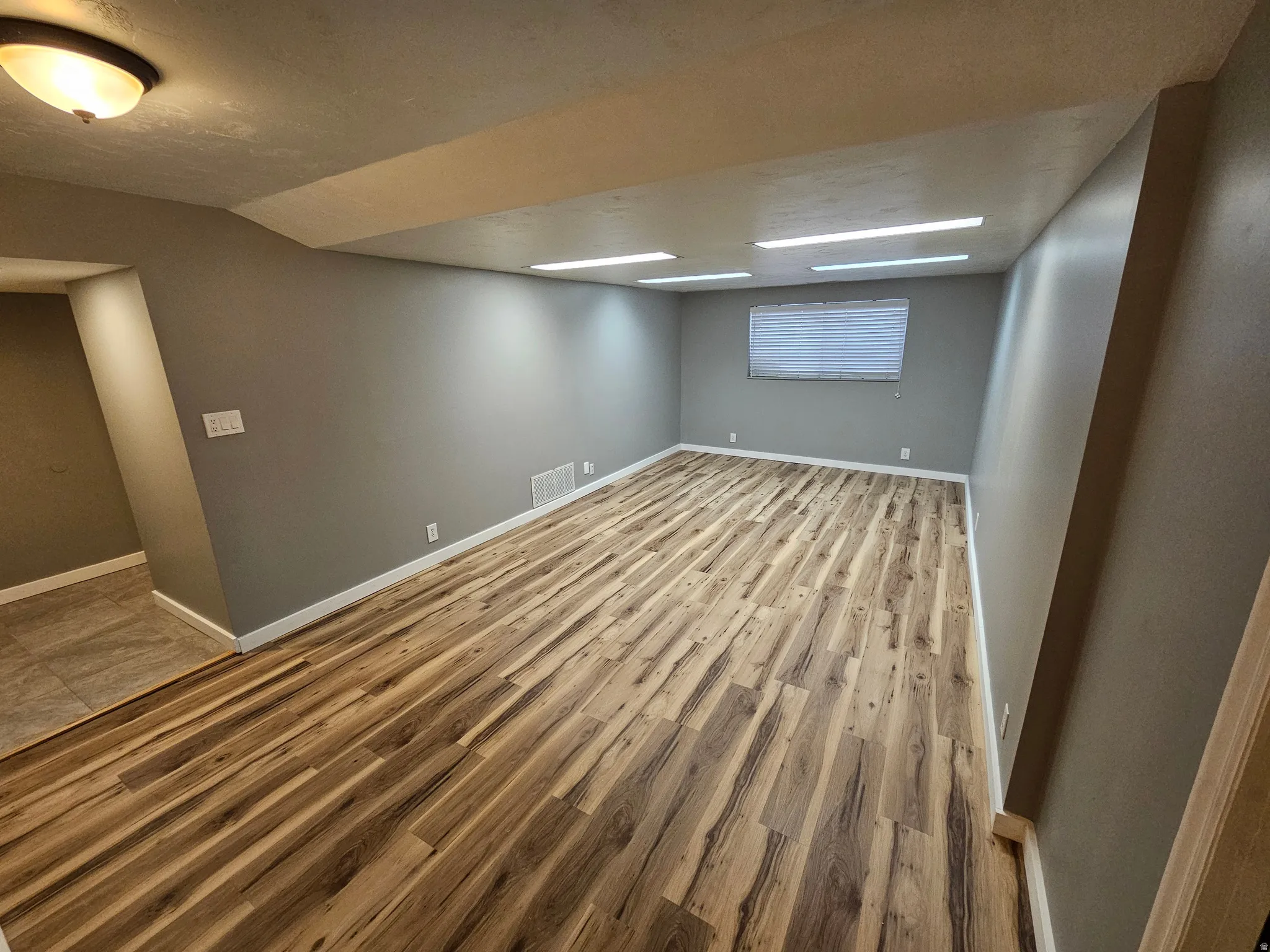 Spacious room featuring wood-finish flooring, neutral gray walls, a window with blinds, and white trim