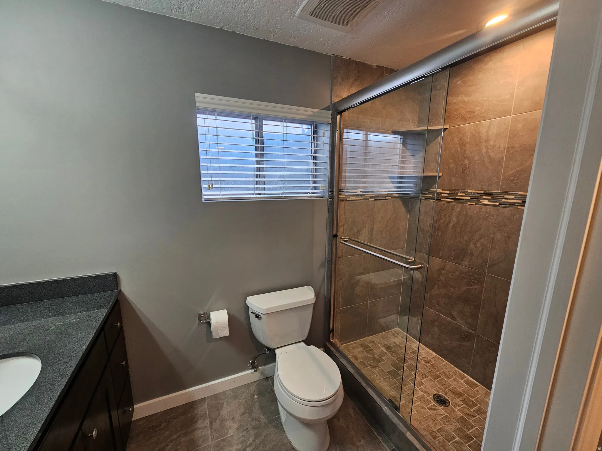 Bathroom featuring a glass-enclosed shower with tile surround, a dark wood-finish vanity with an integrated sink, a dark countertop, and tile flooring